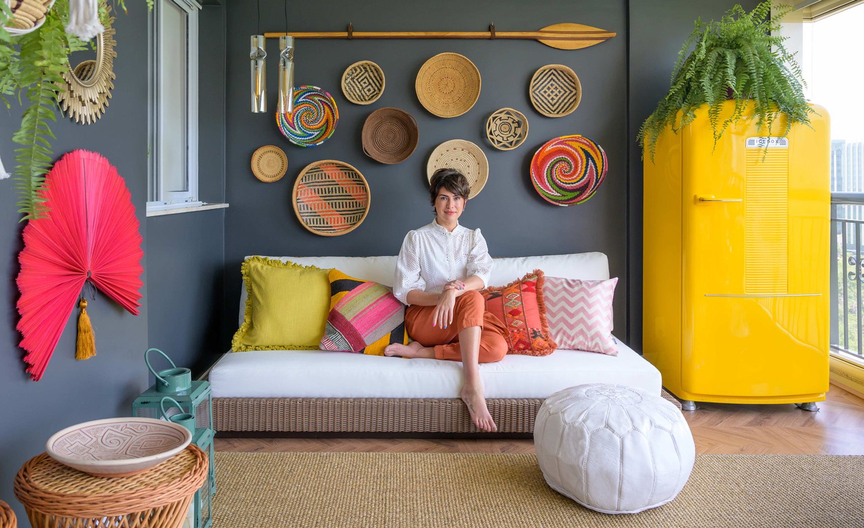 A woman sitting on a white sofa on a colourful balcony decorated with woven baskets and a yellow fridge